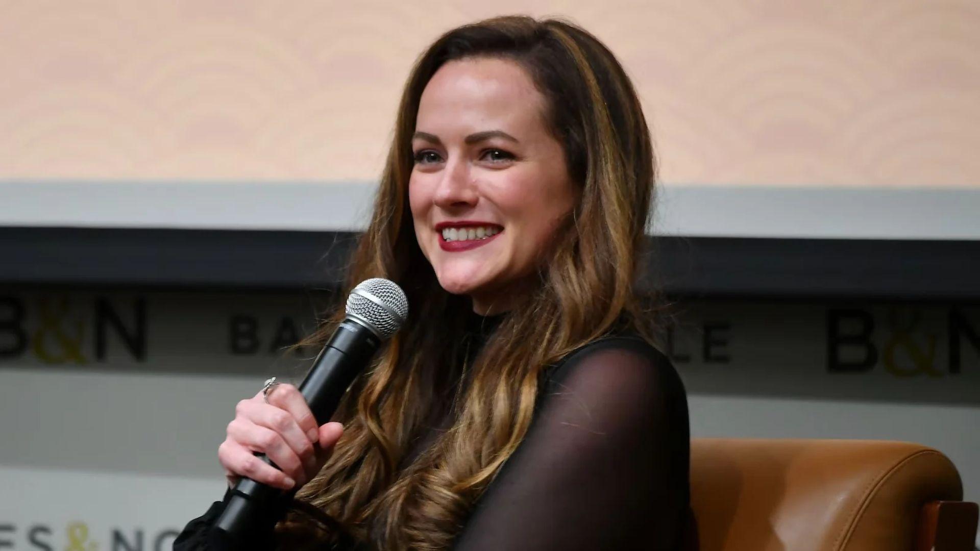 Rebecca Ross smiling while speaking into a microphone at an author event.