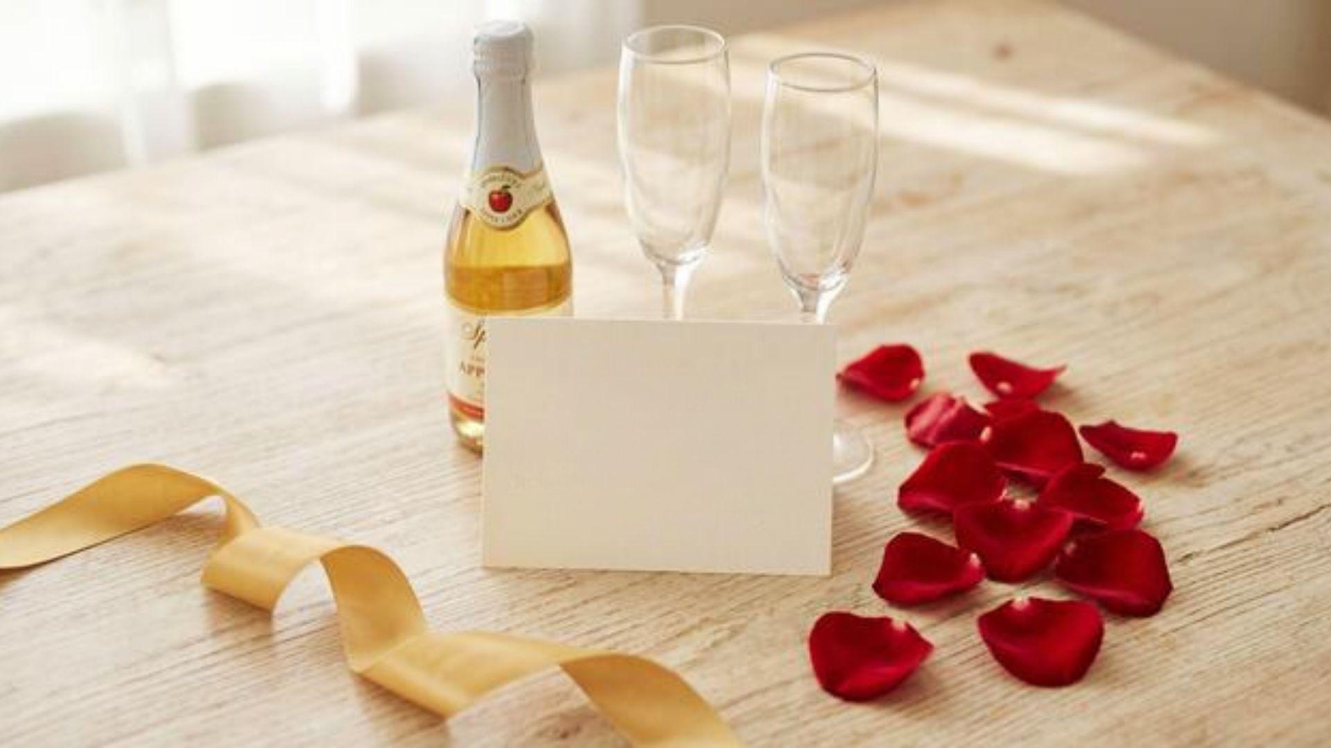 Sparkling bottle, two champagne flutes, blank white card, red rose petals on a table.